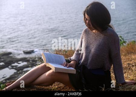 Attractive Asian woman is sitting on the edge of the mountain with a sea view, reading a book. Close-up portrait Stock Photo