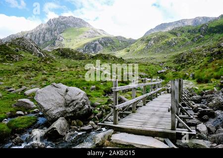 Wooden bridge and footpath crossing stream leading towards mountains Stock Photo