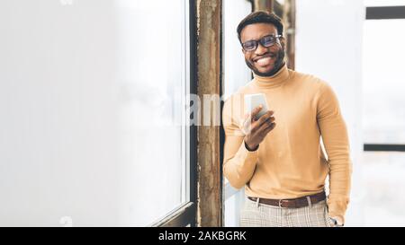 Modern Technologies At Work. Happy african guy using phone in front of window. Panorama, empty space Stock Photo