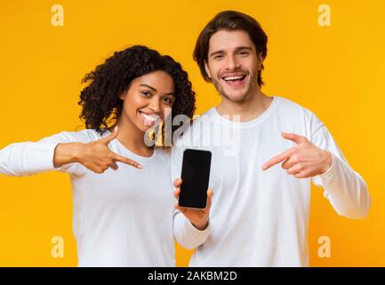 Check this app. Cheerful interracial couple pointing at smartphone with blank black screen for mockup, posing together on yellow background Stock Photo
