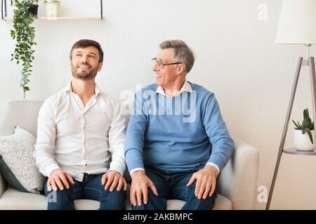 Cheerful Adult Son And Senior Father Talking And Smiling Sitting On Sofa At Home. Relationship With Elderly Parents. Selective Focus Stock Photo