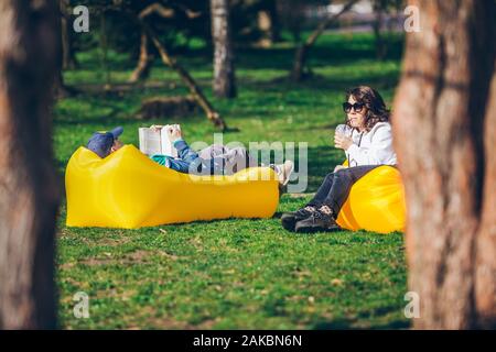couple laying on yellow inflatable mattress in city park. reading book. drinking smoothies Stock Photo