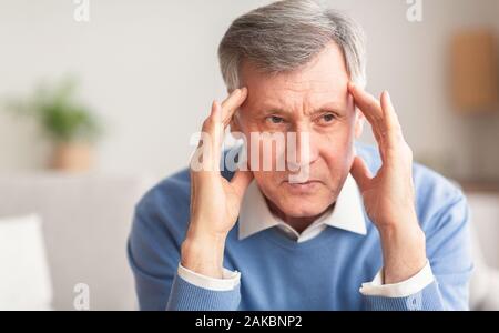 Elderly Man Having Headache Touching Temples Trying To Relieve Pain Sitting On Couch At Home. Selective Focus Stock Photo