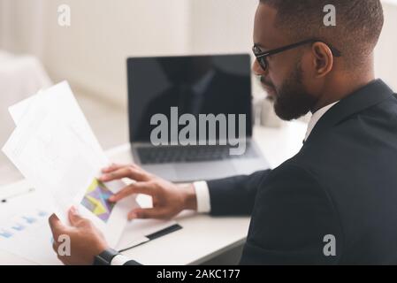 Young black manager in glasses sitting in front of laptop and reading financial report, working in office Stock Photo