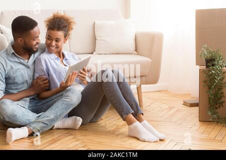 Moving House. Young Afro Spouses Using Digital Tablet Sitting On Floor Among Cardboard Boxes Relaxing In New Flat. Empty Space Stock Photo