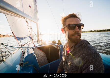 Serious handsome young male yacht captain in sunglasses wearing gloves staying on boat deck and looking into distance. Stock Photo