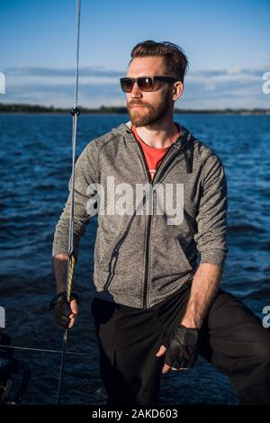 Serious handsome young male yacht captain in sunglasses wearing gloves staying on boat deck and looking into distance. Stock Photo