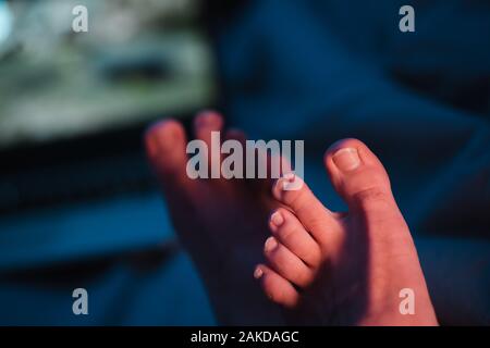 Close up of man toes with long uncut nails that urgently need pedicure - Two feet sitting next to a laptop at night with neon vivid blue red and Stock Photo