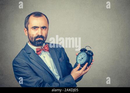 Closeup portrait serious adult bearded man in elegant jacket with red bow tie holding showing alarm clock in his hands looking at you camera, gray wal Stock Photo