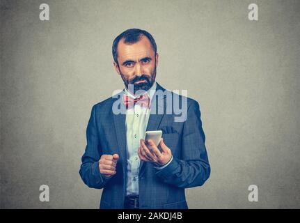 Closeup portrait serious, thoughtful bearded man wearing elegant suit with red bow tie, holding his smartphone in his hand, isolated grey wall backgro Stock Photo