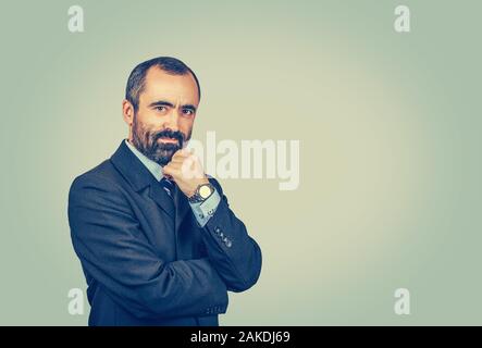 Confident bearded man with dark suit tie blue shirt watch touching chin looking to you camera thinking. Crossed arms. Isolated on light green yellow b Stock Photo