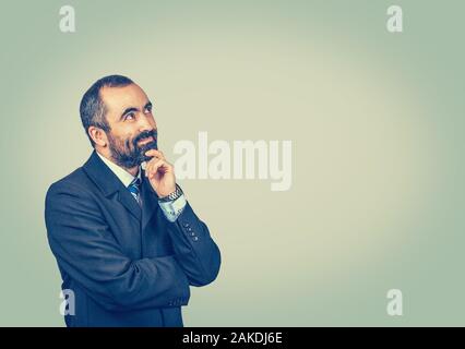 Search idea, thinking. Isolated bearded man with dark suit tie blue shirt watch touching chin, looking up away.  Crossed arms. Isolated bright green y Stock Photo