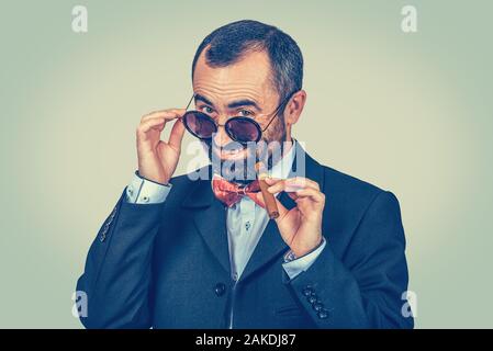 Really? Portrait of smiling, rich, bearded man keeping round sunglasses and cuban cigar. Stylish, funny guy looking you camera. Modern clothes, dark s Stock Photo
