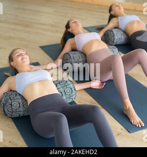 Restorative yoga with a bolster. Group of three young sporty attractive women in yoga studio, lying on bolster cushion, stretching and relaxing during Stock Photo