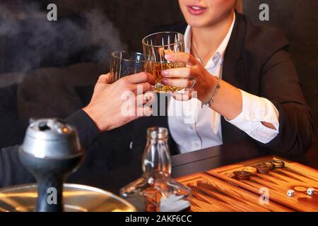 Unrecognizable young woman in white blouse and suit and man drinking whiskey, smoking hookah and playing backgammon in club. Business people resting a Stock Photo