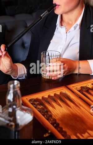 Unrecognizable young woman in white blouse and suit drinking whiskey, smoking hookah and playing backgammon in cafe. Business woman resting after work Stock Photo