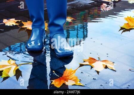 Unrecognizable woman wearing blue rain boots standing in puddle on rainy autumn day. Stock Photo