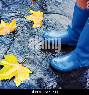Unrecognizable woman wearing blue rain boots standing in puddle on rainy autumn day. Stock Photo