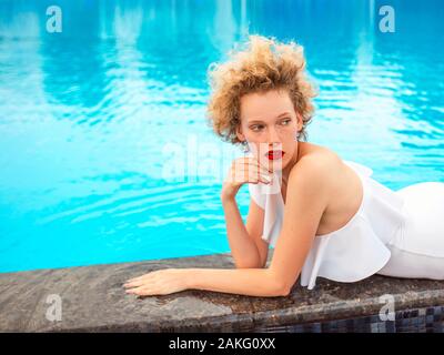 beautiful redhead (ginger) curly woman in white swimming suit laying by the swimming pool in summer day. Summer, happiness, relax, wellness concept Stock Photo