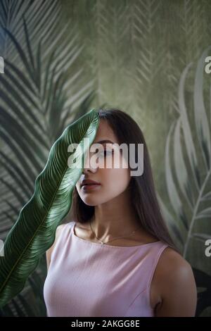 Gentle portrait of a beautiful young woman with a leaf of a plant on her face. Tropical background. Stock Photo