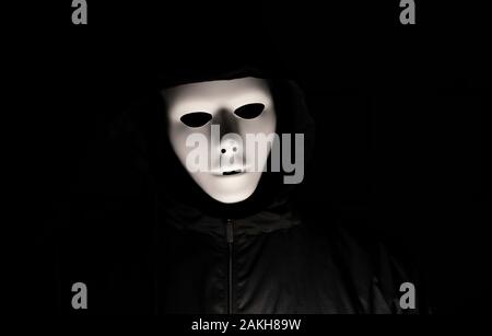 Scary white mask in the dark. Spooky guy in black jacket and white masquerade. Black, invisible eyes. Halloween style. Stock Photo