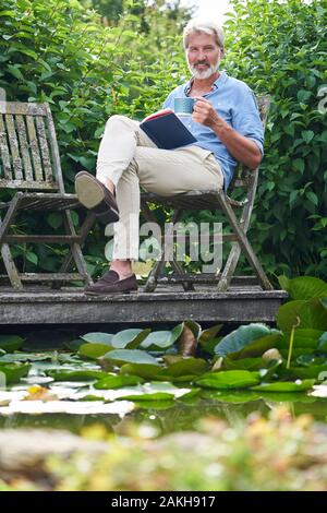 Portrait Of Mature Man Relaxing In Garden Reading Book On Jetty By Lake Stock Photo