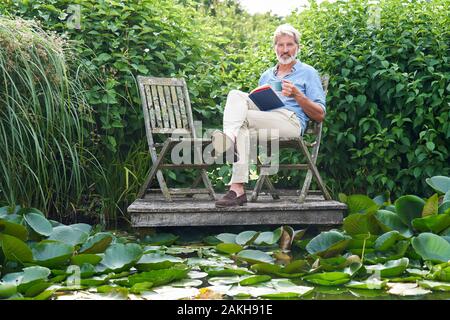 Portrait Of Mature Man Relaxing In Garden Reading Book On Jetty By Lake Stock Photo