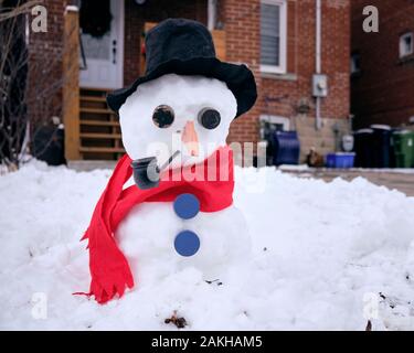 Snowman dressed with red scarf black felt hat blue buttons smoking a pipe in front of Leaside residence Stock Photo