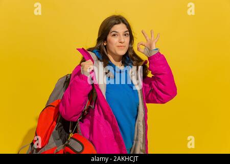 Smiling, shows nice. Portrait of a cheerful young caucasian tourist girl with bag and binoculars isolated on yellow studio background. Preparing for traveling. Resort, human emotions, vacation. Stock Photo