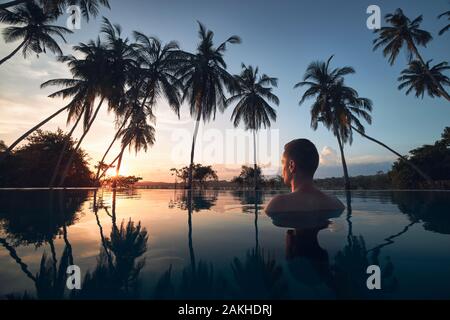 Young man watching sunset from swimming pool in the middle of coconut palm trees.