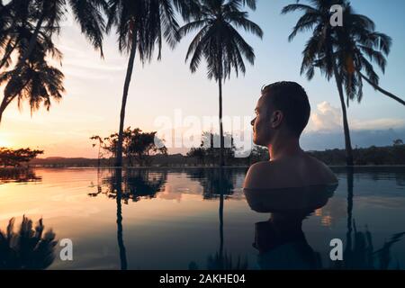 Young man watching sunset from swimming pool in the middle of coconut palm trees.
