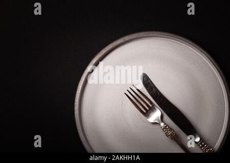 Closeup empty round gray ceramic plate on black background, vintage cupronickel fork, knife with copy space. Concept modern shooting menu from chef, r Stock Photo