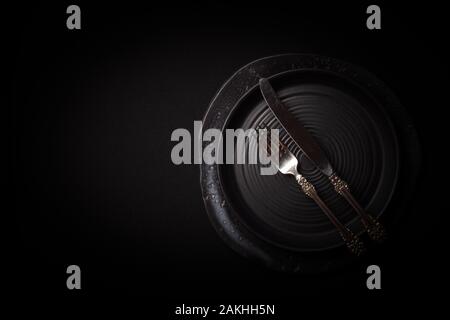 Closeup 2 empty round black aged ceramic plates, vintage cutlery, silver fork, knife on dark background with copy space. Concept modern shooting menu, Stock Photo