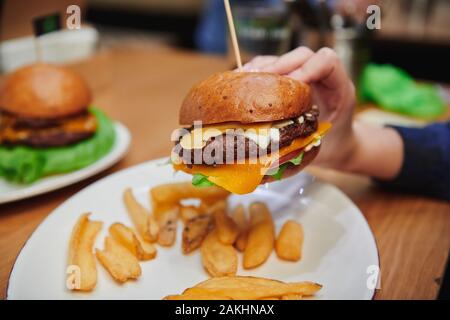 Juicy cheeseburger in girls hand. Natural beef burger with double cheese served with potato dippers. High-calorie dinner in the cafe. Stock Photo