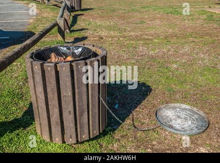 In a park at the parking lot is an overflowing garbage can with plenty of trash inside with the lid laying on the ground on a bright sunny day Stock Photo