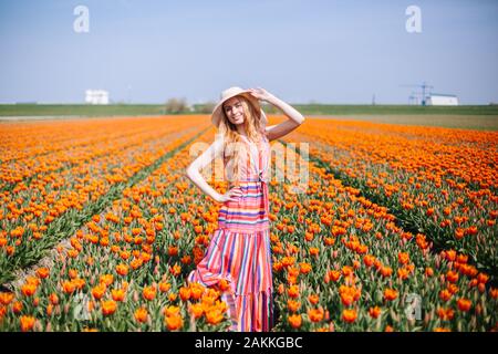 Magical landscape with beautiful young long red hair woman wearing on striped dress and straw hat standing on colorful flower tulip field in Holland. Stock Photo