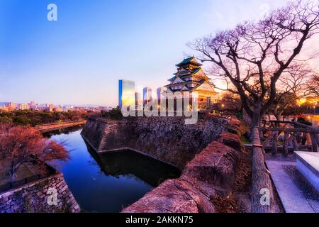 The main keep of Osaka castle from shogun times rising over stone walls and water filled moats at sunrise against clear sky and distant modern busines Stock Photo