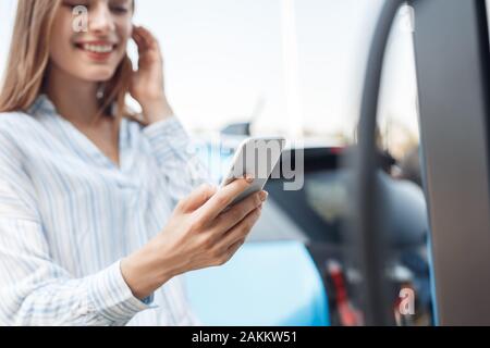 Transportation. Young woman on electric car having stop at charging station using app on smartphone close-up blurred background cheerful while getting Stock Photo