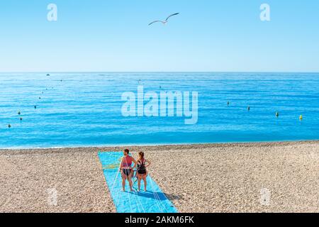 A young couple waits for a boat to being parasailing on the pebbly beach of the Bay of Angels on the French Riviera of the Mediterranean Sea. Stock Photo