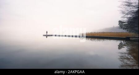 Man standing alone on edge of pier and staring at lake. Mist over water. Foggy air. Early chilly morning in autumn. Beautiful freedom moment and peace Stock Photo