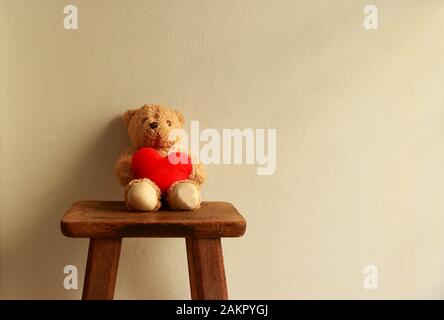 Closeup bear doll holding red heart sitting on vintage wooden chair on sunshine day with cement wall background, love concept Stock Photo