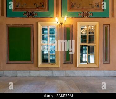 Two narrow wooden window with closed green shutters and beautiful elegant rectangular green frames on orange wall with white marble floor Stock Photo