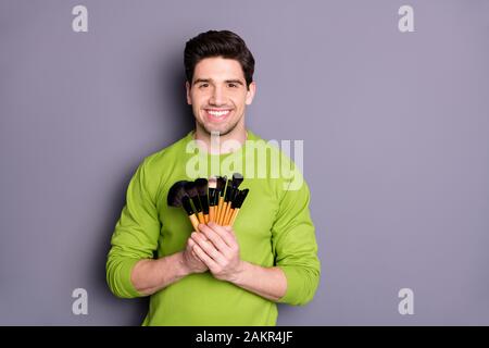 Portrait of positive successful man visagiste hold brushes to make maquillage for special evening events photo session wear green sweater isolated Stock Photo