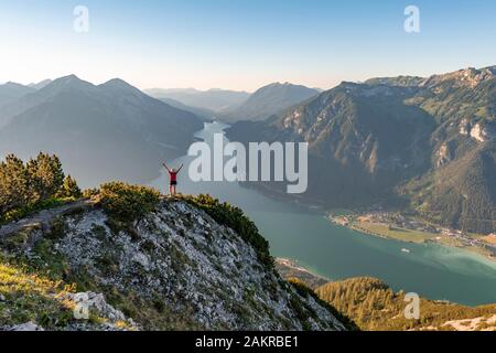 Young woman stretches arms in the air, view from the mountain Baerenkopf to Lake Achensee, left Seebergspitze and Seekarspitze, right Rofan Stock Photo