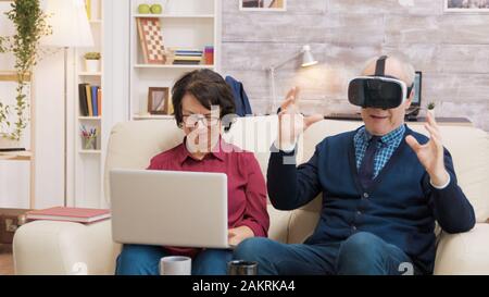 Senior man trying a VR headset in the living room while his wife uses a laptop next to him. Modern old couple using technology Stock Photo