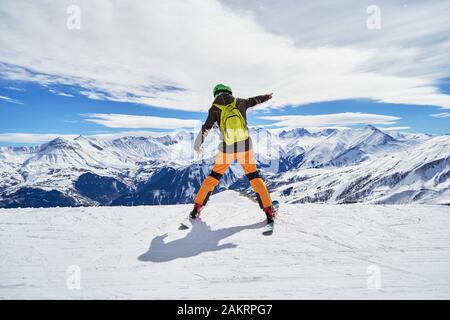 Enthusiastic skier wearing colorful clothes and a green backpack, posing on a ski slope in Les Sybelles ski resort, with French Alps peaks in the back Stock Photo