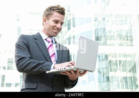 Smiling businessman standing outdoors typing on his laptop in front of background of modern glass office towers Stock Photo