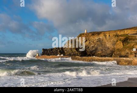 Stormy weather at Portreath, Cornwall, UK with waves breaking over the Monkey Hut and the Pepperpot visible on the high cliff. Stock Photo