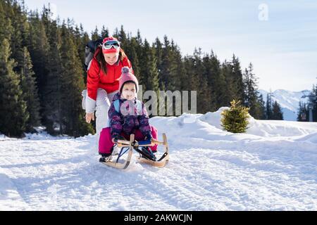 Adorable Girl Enjoying Sledding While Her Mother Pushing Sledge Against Pine Tree Forest During Winter Stock Photo