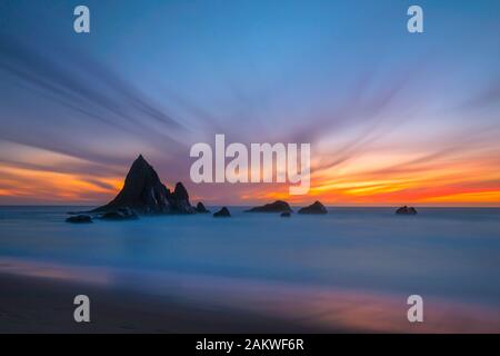 A colorful sunset backdrops the unique rock formations of Shark Fin Rock on Martin's Beach, in Half Moon Bay, California as the waves come in Stock Photo